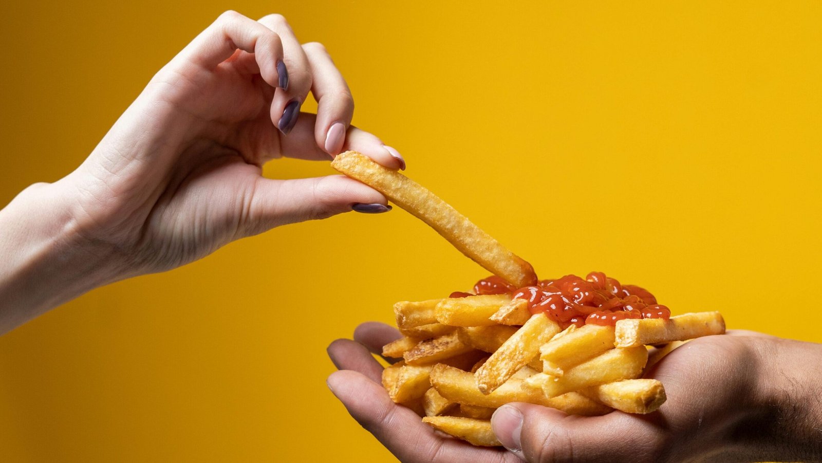 pexels-photo-4676401-4676401 A close-up of hands sharing a delicious pile of french fries with ketchup against a yellow background.